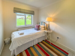 Single bedroom at Penamont with bedside table, lamp and rug on painted wooden floor. Window to the rear garden