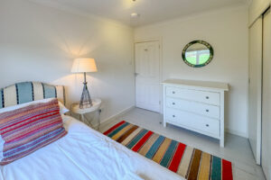 Single bedroom at Penamont with bedside table, lamp and rug on painted wooden floor. Chest of drawers with mirror over, built in wardrobe and door to hallway