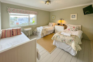 Triple bedroom at Penamont with 3 single beds, window to the front of the house, chest of drawers with lamp in between two single beds, Painted floor with rug in between single beds and wall mounted TV in corner