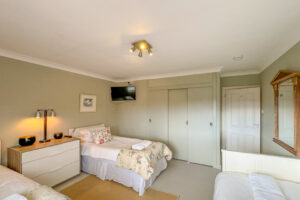 Triple bedroom at Penamont with 3 single beds, chest of drawers with lamp in between two single beds, Painted floor with rug in between single beds built in wardrobe and wall mounted TV in corner